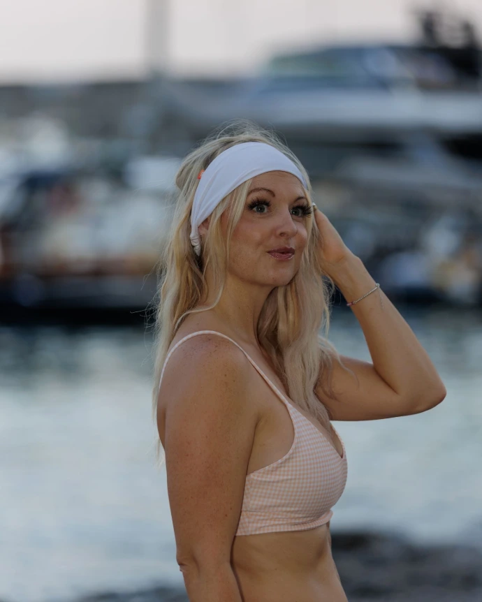 Blonde model photographed in profile in front of a softly blurred harbour, hand raised to her hair, wearing the Bikiboom Wide Headband in White sitting wide and flat across the forehead. Paired with a blush pink gingham bikini top, the white wide headband demonstrates how this elasticated headband works as a versatile hair accessory that coordinates effortlessly with any colorway. Soft evening light, natural editorial mood. The image highlights the Bikiboom Wide Headband in White as the essential summer headband and beach headband for headband women who want a clean, minimal hair band that goes with everything.
