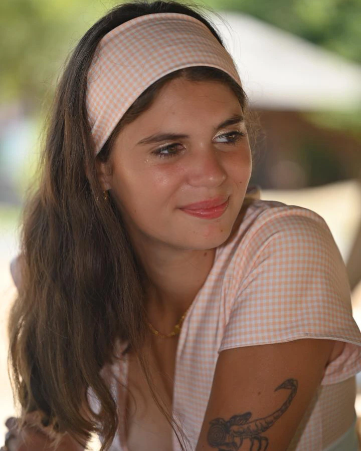 Close-up portrait of a young woman wearing the Sunfire Mirage elasticated headband in pastel orange and white gingham, hair worn naturally down. Soft, warm light with a blurred green background. The wide shape of the hair band sits flat and structured across the hairline, framing the face cleanly. A relaxed, joyful expression that captures the effortless beach headband energy of the Sunfire Mirage colorway.