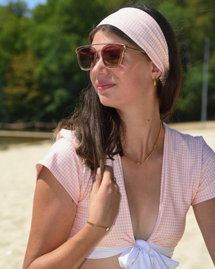 Young woman on a sandy beach wearing a wide headband in the Sunfire Mirage pastel orange and white gingham print, paired with a matching gingham top tied at the waist over a white bikini bottom. Gold chain necklace and bracelet, oversized tinted sunglasses. Hair worn down, face turned to the side in natural sunlight. A confident, effortless summer look that shows the headband as a full outfit accessory, not just a hair accessory.